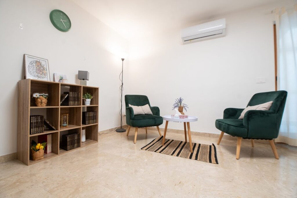 Arredamento di uno studio con due poltrone in velluto verde scuro, un tavolino da caffè bianco con decorazioni floreali, un piccolo tappeto a righe e una libreria in legno con libri e soprammobili.